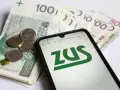 Złotówki i monety obok telefonu z logo ZUS. Pamiętaj o płaceniu składek, bo kto nie płaci funduszu pracy, ten ma problemy.