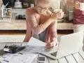 Kobieta w okularach analizuje dokumenty przy laptopie, obok kalkulator i kawa.