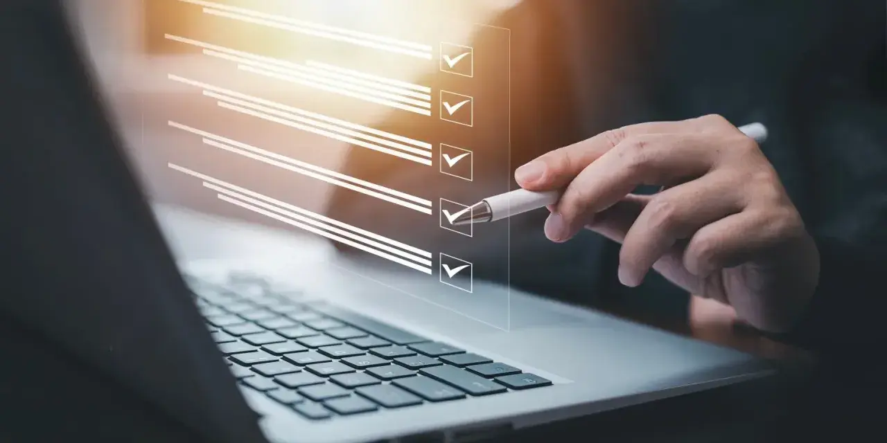 Ręka z długopisem zaznacza punkty na liście kontrolnej na ekranie laptopa. Proces ten może pomóc w ustaleniu, jak zmienić regulamin zakładowego funduszu świadczeń socjalnych.