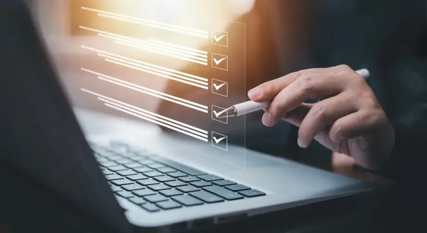 Ręka z długopisem zaznacza punkty na liście kontrolnej na ekranie laptopa. Proces ten może pomóc w ustaleniu, jak zmienić regulamin zakładowego funduszu świadczeń socjalnych.