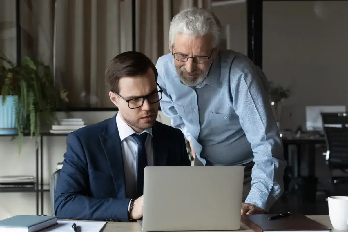 Dwóch mężczyzn analizuje dane na laptopie. Starszy mężczyzna pochyla się nad młodszym, dyskutując o tym, kto nie płaci funduszu pracy.